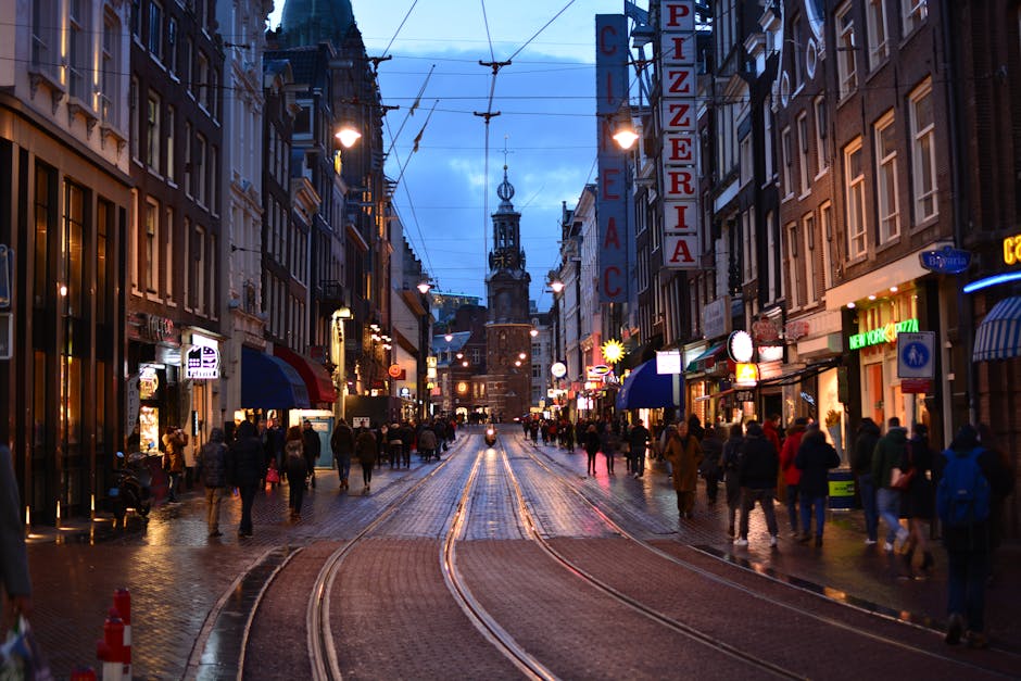 Cena de rua dinâmica ao entardecer em Amesterdão, com multidões animadas e lojas iluminadas.