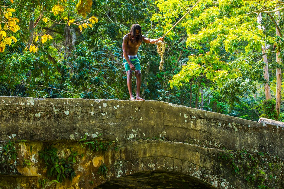 Um homem afro-americano está numa antiga ponte de pedra numa floresta luxuriante da Jamaica, a segurar uma corda.