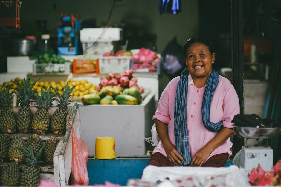 Mulher sorridente sentada numa banca colorida de mercado de fruta com ananases e outras frutas.