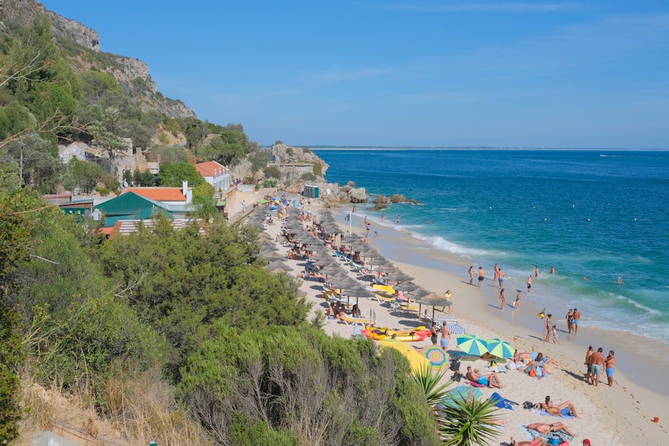 Um dia de verão movimentado na praia do Portinho da Arrábida, com banhistas e guarda-sóis ao longo da costa.
