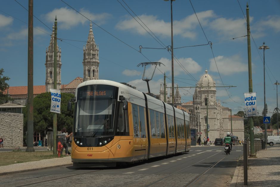 Um elétrico amarelo passa pelo icónico Mosteiro dos Jerónimos em Lisboa, sob um céu azul límpido.