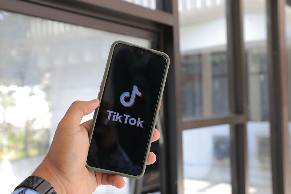 Close-up de uma mão a segurar um smartphone com a aplicação TikTok aberta, em ambiente interior.