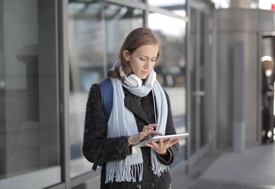 Jovem mulher em pé no exterior, a usar um tablet, com auscultadores e um cachecol, representando tecnologia e viagens.