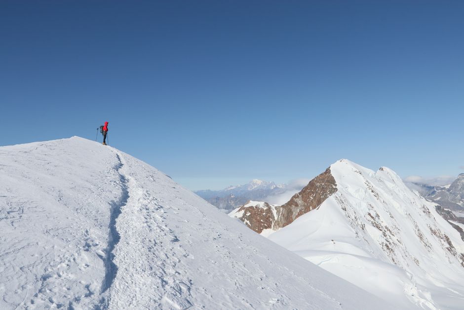 Um alpinista solitário no cume de um pico coberto de neve, sob um céu de inverno límpido, a demonstrar a aventura do trekking em montanha.