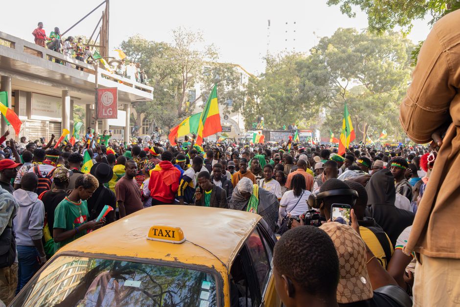 Uma cena de rua animada em Dakar, Senegal, com uma multidão a celebrar e a acenar bandeiras.