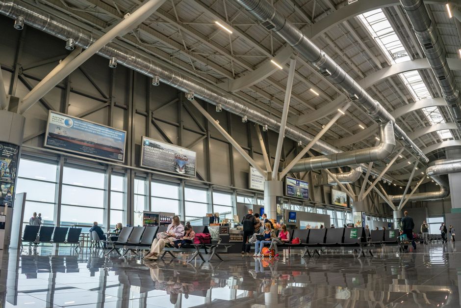 Interior de um terminal de aeroporto espaçoso com passageiros sentados e a caminhar, exibindo um design moderno e janelas amplas.