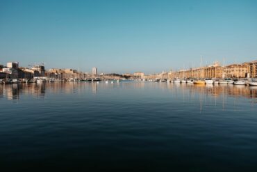 Vista refletida de veleiros no porto de Marselha sob um céu azul límpido.