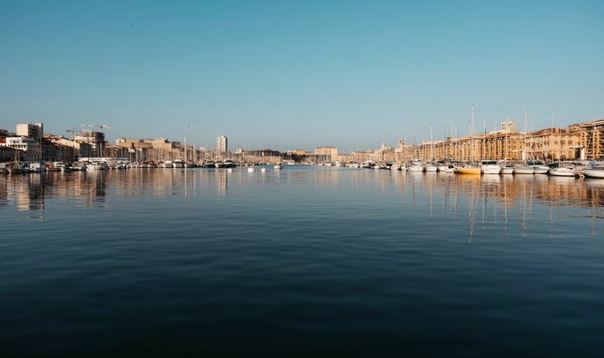 Vista refletida de veleiros no porto de Marselha sob um céu azul límpido.