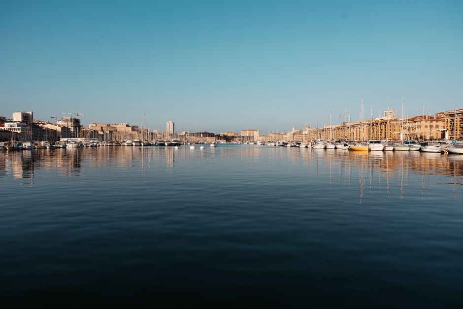 Vista refletida de veleiros no porto de Marselha sob um céu azul límpido.